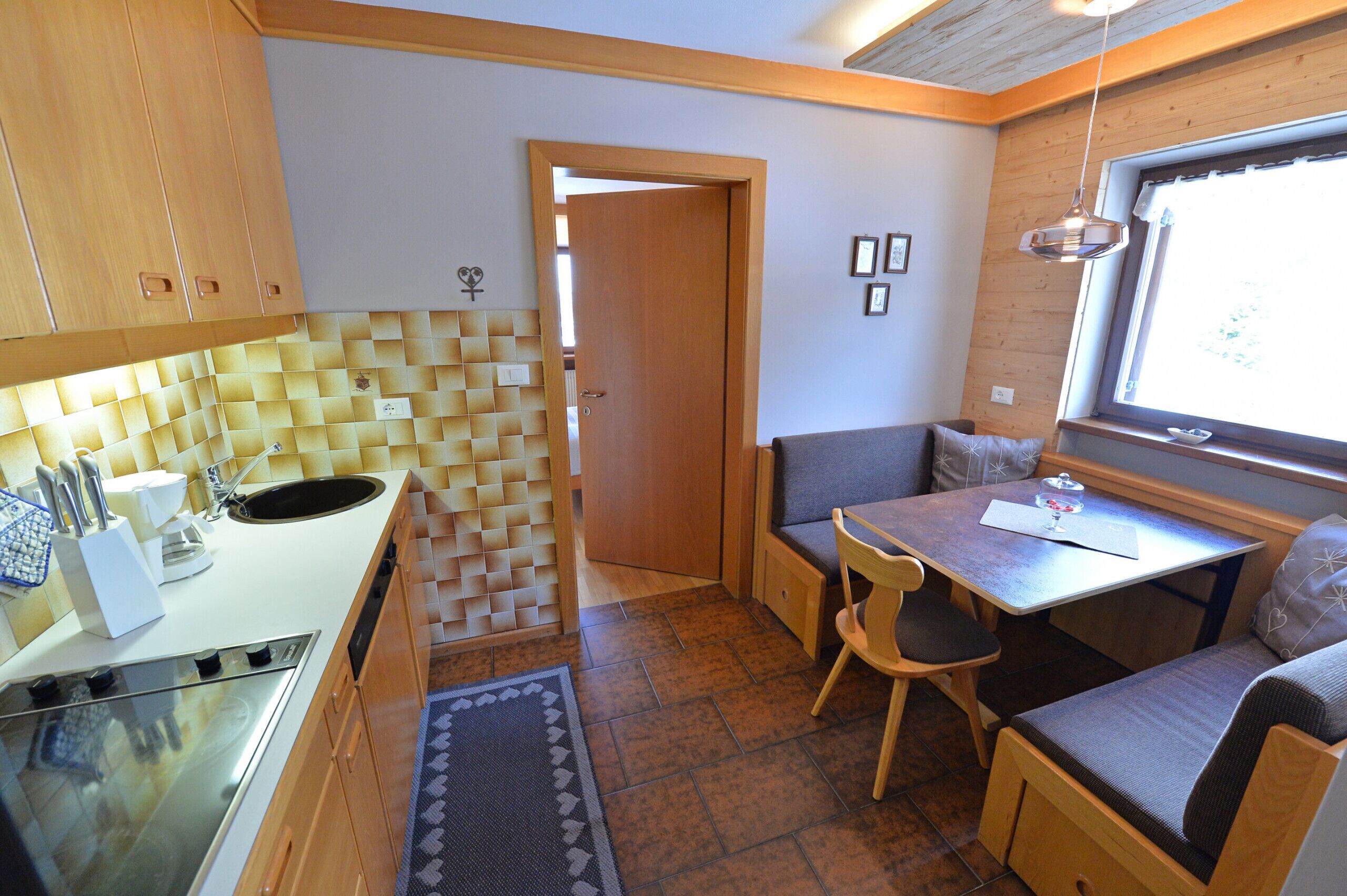 Una cucina compatta con lavello, fornello e mobili in legno. C'è un'area pranzo con tavolo e sedie, e una panca con cuscini.