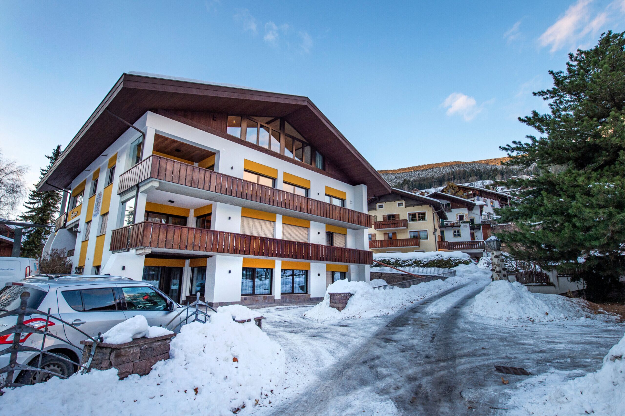 Un hotel a più piani con balconi, un vialetto coperto di neve e montagne sullo sfondo.