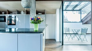 Een moderne keuken met een centraal eiland, een hangende kristallen kroonluchter en een uitzicht op een zitgedeelte buiten door een glazen deur.