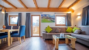 A cozy living room with a wooden ceiling, a sofa with cushions, a glass coffee table, and a dining area with a table and chairs.