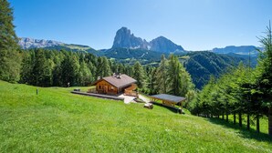 A wooden cabin with a solar panel, surrounded by lush greenery and mountains.