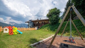 A playground with a swing set and a slide, surrounded by a wooden fence and a building with a balcony. - Residence Chalet Alpina 143E (Ortisei)