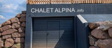 Rakennuksessa on kyltti, jossa lukee ”CHALET ALPINA.info”, ja sen yläpuolella on suljettu autotallin ovi.