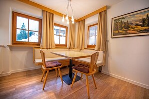 A dining area with a wooden table and chairs, large windows with curtains, and a painting on the wall. - Residence Chalet Alpina 143C (Ortisei)