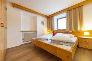 A wooden bed with white linens, a bedside table with a lamp, a window with curtains, and a radiator. - Residence Chalet Alpina 143C (Ortisei)