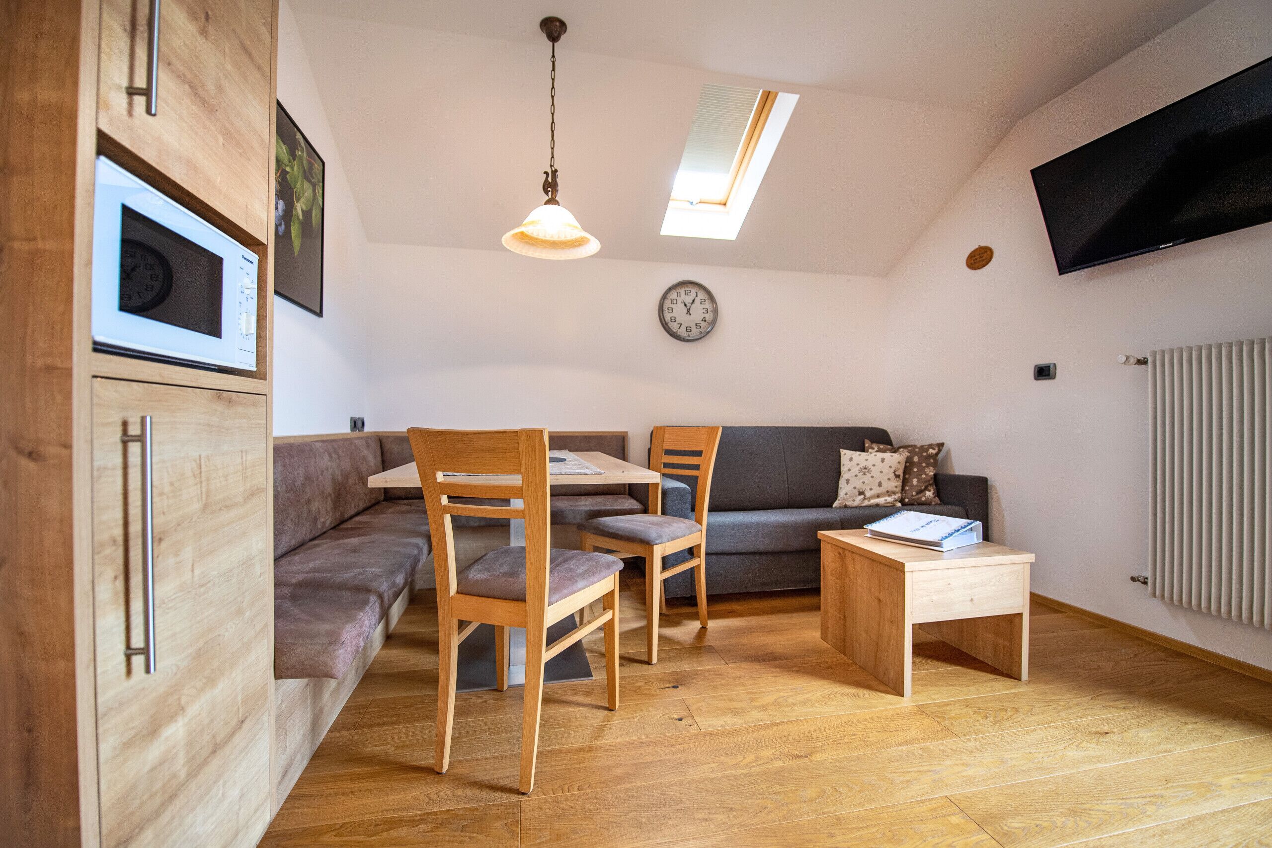 A compact living space with a dining area, a sofa, a TV mounted on the wall, and a clock.