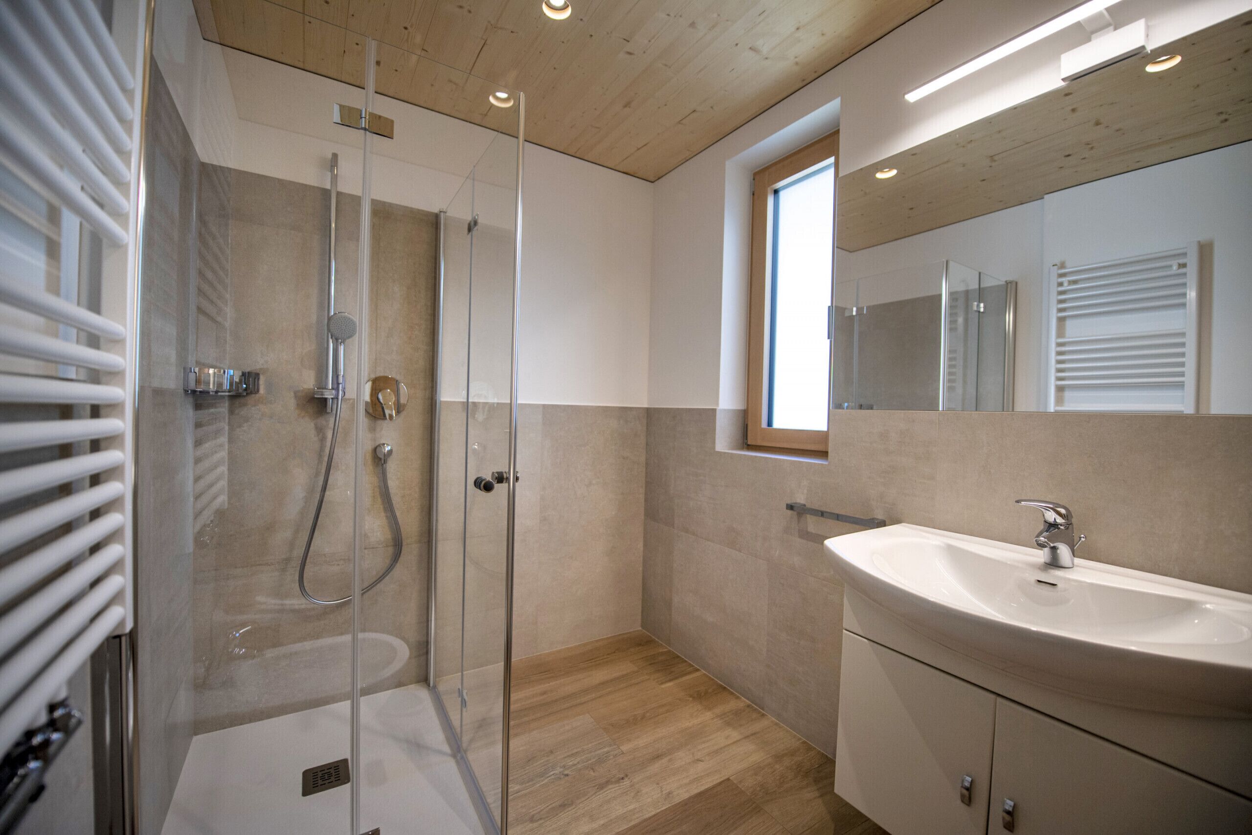 Ein modernes Badezimmer mit einer gläsernen Duschkabine, Holzboden, großem Spiegel und weißem Waschbecken.