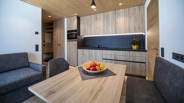 Een moderne keuken met houten kastjes, een inbouwoven en een aanrecht met een plant in pot. Er is een eettafel met een fruitschaal en een grijze bank in de woonkamer.