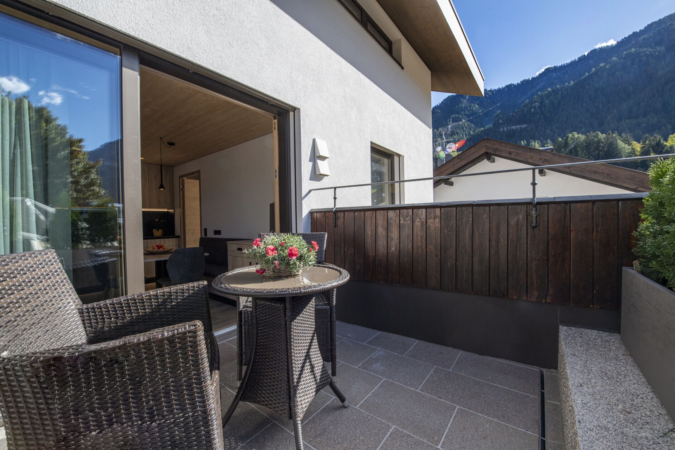 A patio with wicker furniture and a table with flowers, a wooden fence, and a mountain view.