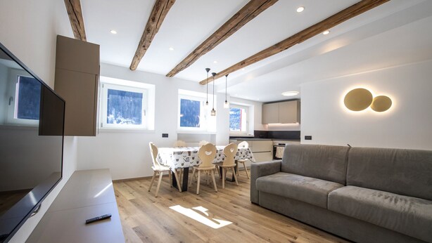 A modern living room with a wooden ceiling, a grey sofa, a dining table with chairs, and a TV on a stand.