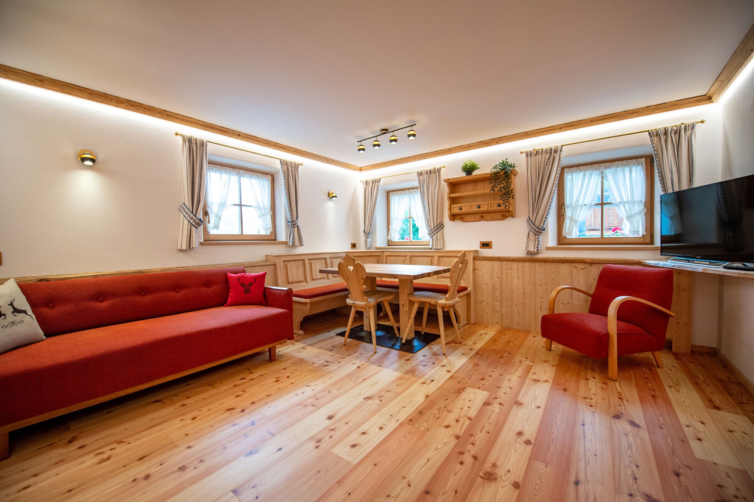 A room with a red sofa, wooden dining table, and chairs. There is a TV on the right wall and windows with curtains.