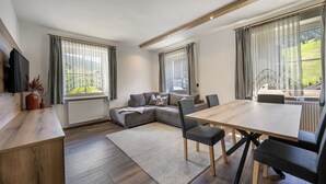 Ein modernes Wohnzimmer mit Sofa, Esstisch und Stühlen, großen Fenstern mit Vorhängen und einem Holzbalken an der Decke.
