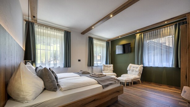 Ein Schlafzimmer mit einem großen Bett, zwei Stühlen, einem Fernseher und einem Fenster mit Vorhängen.