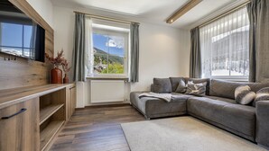 Ein modernes Wohnzimmer mit einer Eckcouch, Holzschränken, einem Flachbildfernseher und einem großen Fenster mit Bergblick.