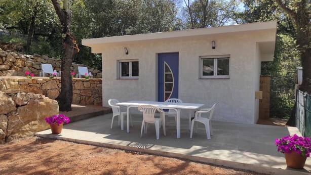 A small, single-story building with a white exterior and a blue door. It features a concrete patio with a white table and chairs, surrounded by stone walls and potted plants. - Country cottage (Flassans-sur-Issole)