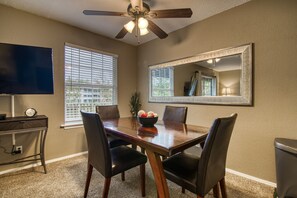 A dining area with a wooden table, four chairs, a ceiling fan, a large mirror, and a TV on a stand.