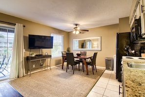 A compact kitchen with a dining area, a ceiling fan, a TV, and a large window with curtains.