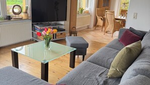 A living room with a grey sofa, a glass coffee table, and a flat-screen TV. There are wicker chairs and a dining table in the background.