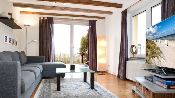 A modern living room with a grey sofa, a glass coffee table, wooden flooring, and a large window with curtains.