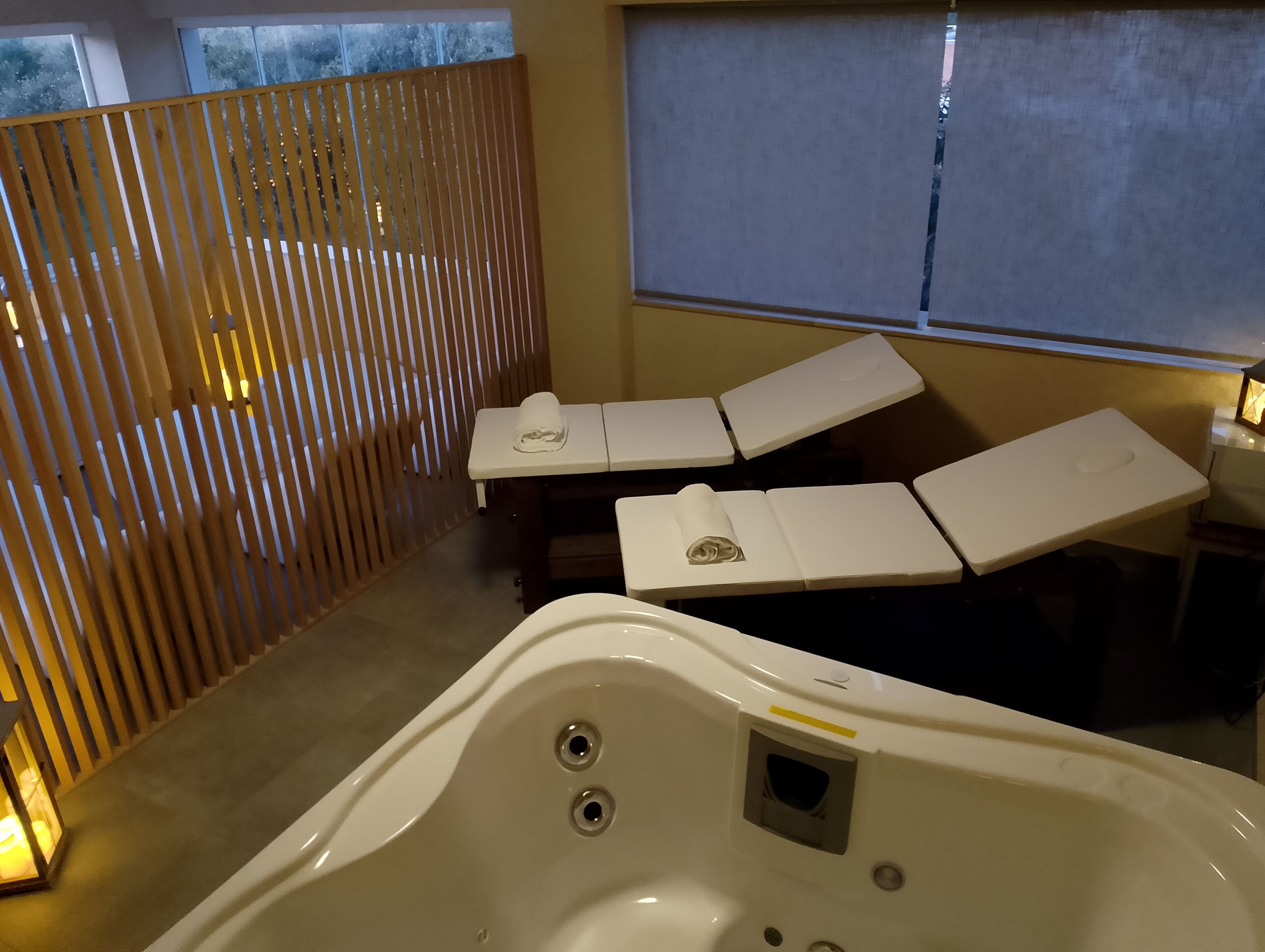 Un espace bien-être comprenant une baignoire, deux chaises longues, une cloison en bois et une fenêtre avec des stores.
