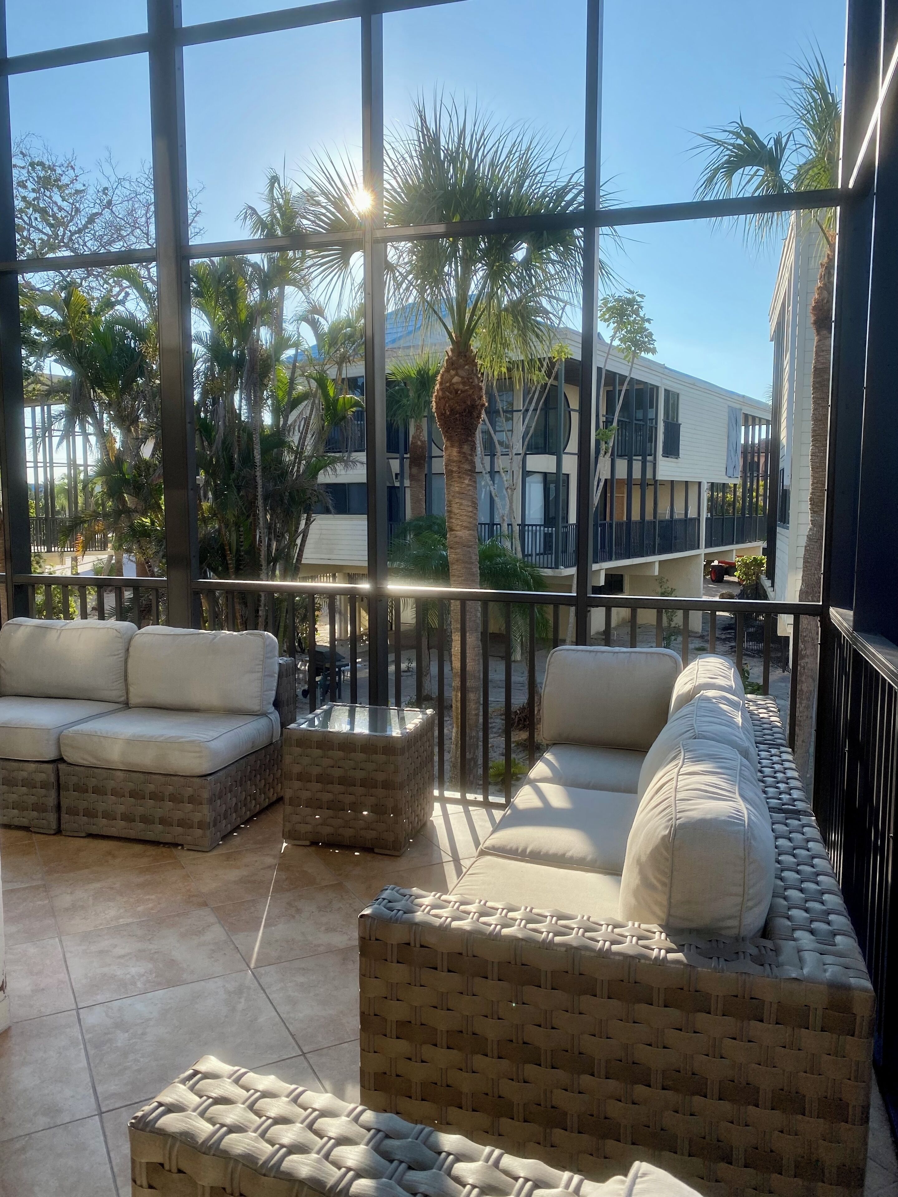 A sunlit outdoor seating area with wicker furniture and a glass-top table, overlooking a building and palm trees.