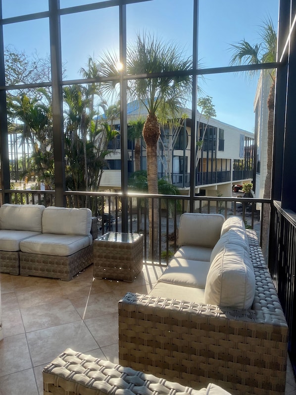 A sunlit outdoor seating area with wicker furniture and a glass-top table, overlooking a building and palm trees.