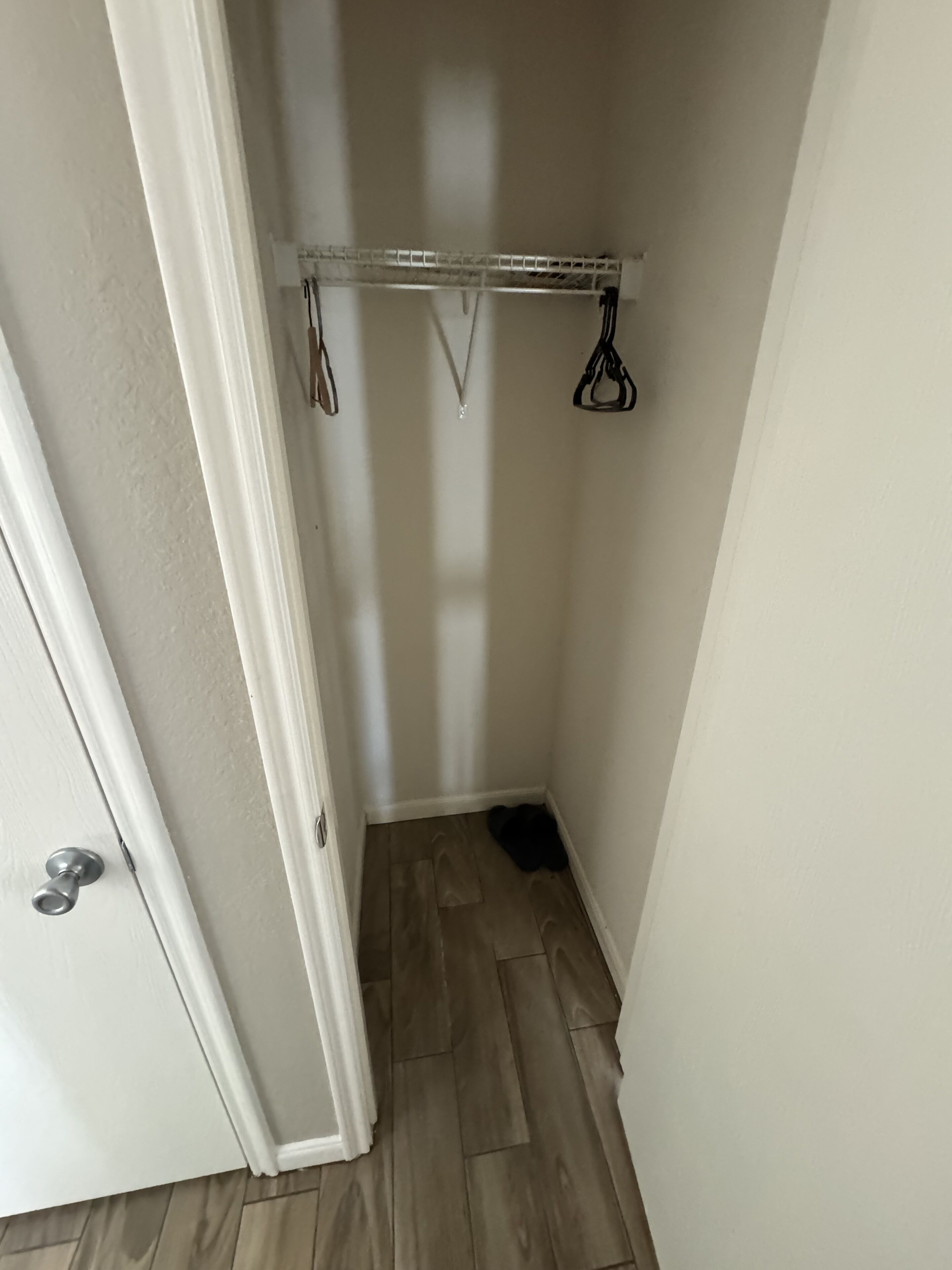 A small, narrow hallway with a wooden floor and a white door on the left. A metal rack with hooks is mounted on the wall, holding a few items.