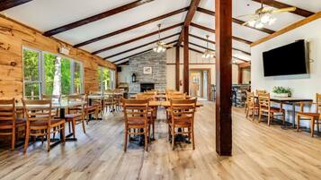 A spacious room with wooden floors and tables, large windows, a stone fireplace, and a television on the wall.