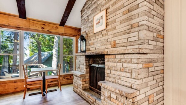 A room with a stone fireplace, wooden beams, a dining table with chairs, and large windows. - Rare Muskoka Chalet Suite - B8 (Dwight)