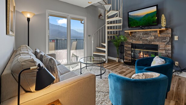 Un salon moderne avec un canapé, des fauteuils bleus, une cheminée, un téléviseur, un ventilateur de plafond et un escalier.