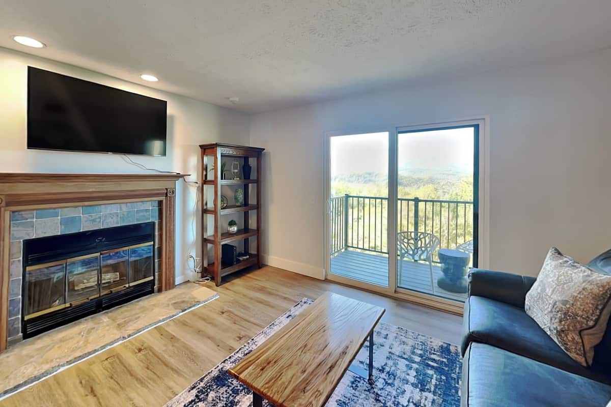 A living room with a fireplace, a TV mounted on the wall, a bookshelf, and a balcony with a view of the outdoors.