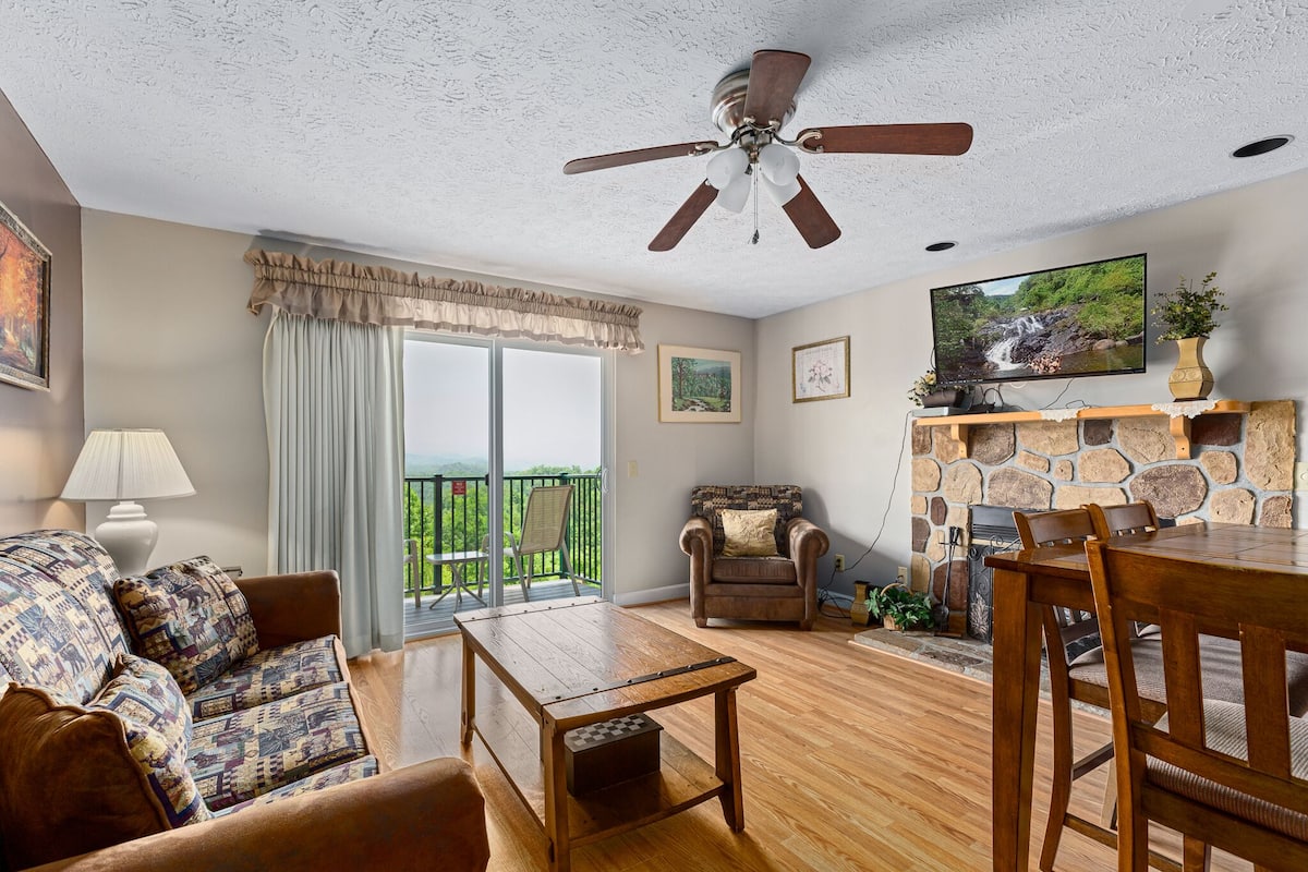 A living room with a sofa, a coffee table, a dining table, a fireplace, a ceiling fan, and a balcony with a view.