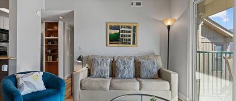 A living room with a blue armchair, a beige sofa with patterned cushions, a glass coffee table, a ceiling fan, a framed landscape painting, and a large window with blinds.