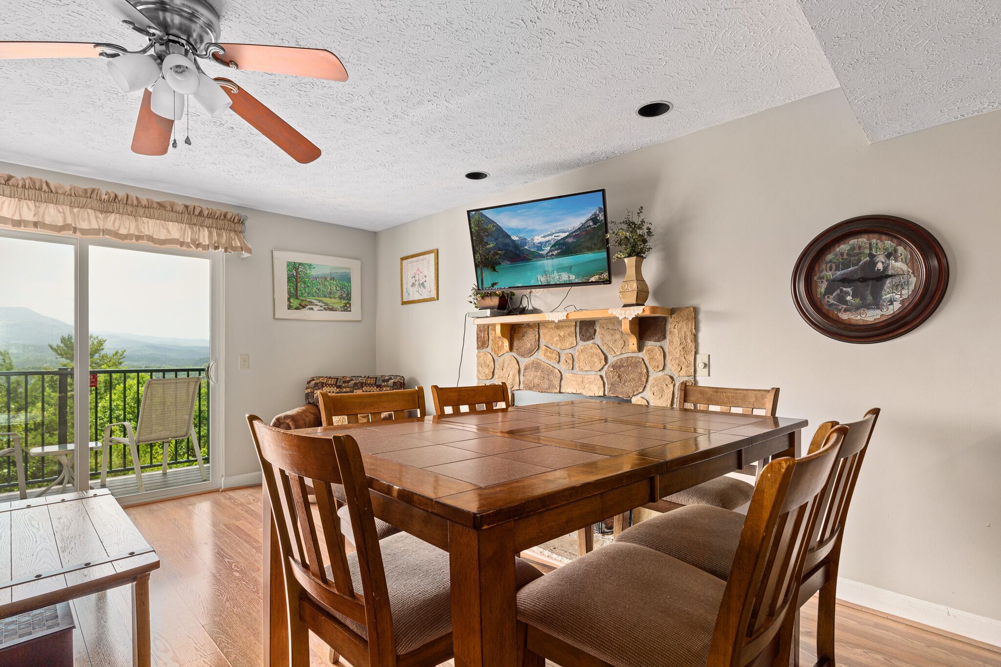 A dining area with a wooden table and chairs, a ceiling fan, a television, and a stone fireplace.