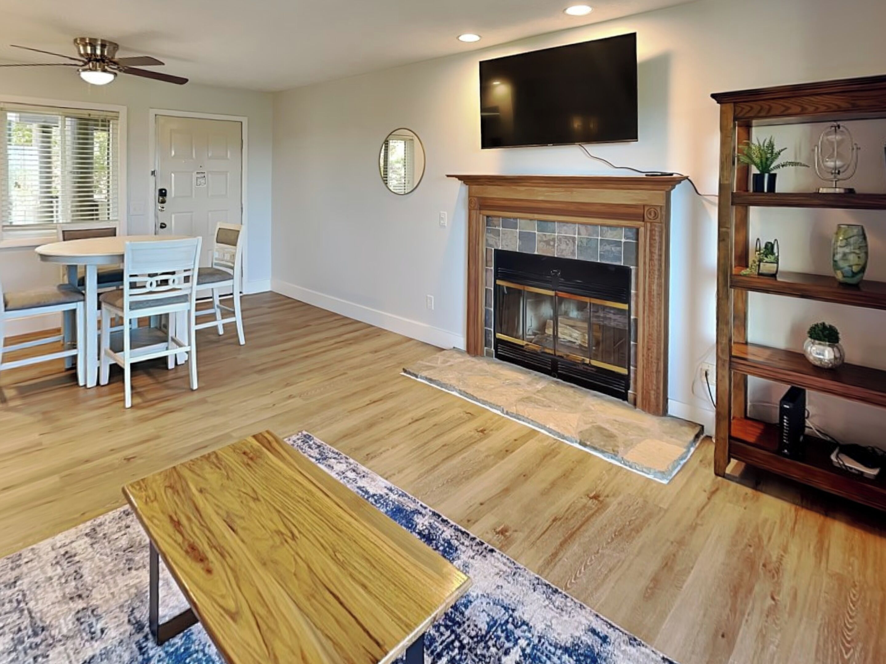 A living room with a fireplace, a television mounted above it, a dining area with a table and chairs, and a bookshelf with decorative items.