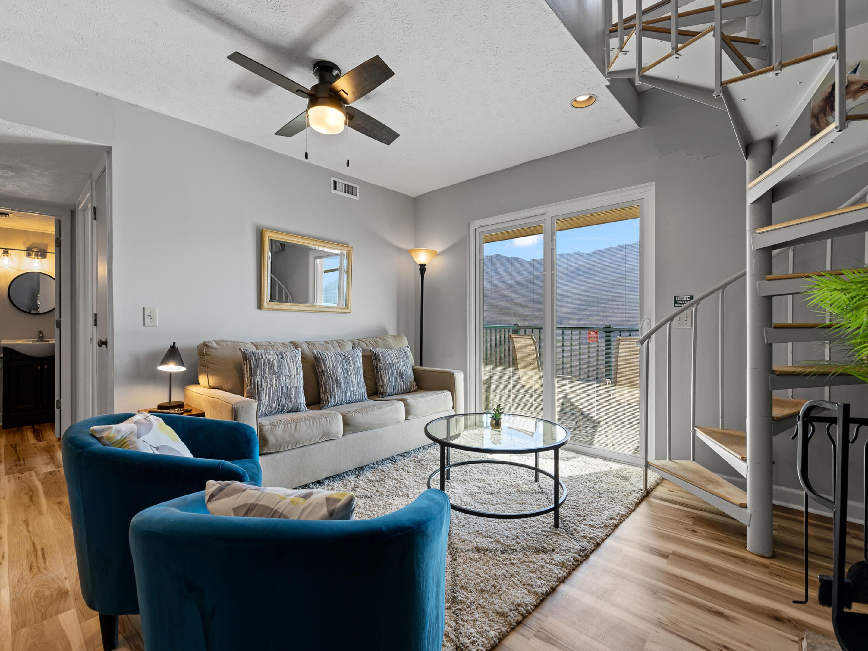 Un salon moderne avec un ventilateur de plafond, un canapé agrémenté de coussins à motifs, une table basse et un escalier offrant une vue dégagée.