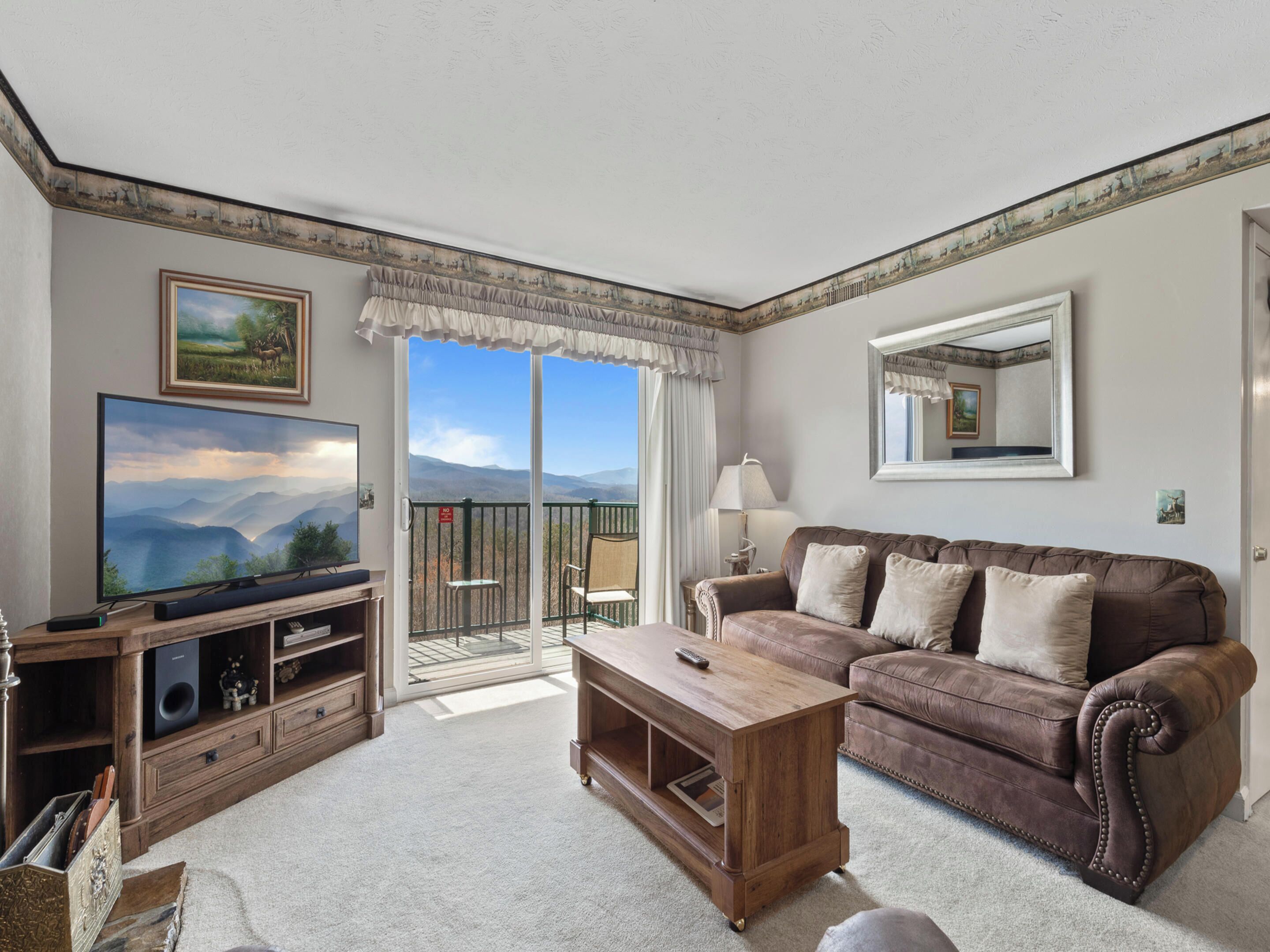 A living room with a television, a brown leather sofa, a wooden coffee table, and a balcony with a view of mountains.