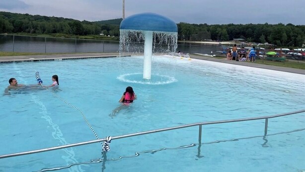 A swimming pool with a water fountain feature, surrounded by a fence, with people swimming and standing by the pool. - Villa in Arrowhead Pocono Resort community Walking to the Lake  . (Pocono Lake)
