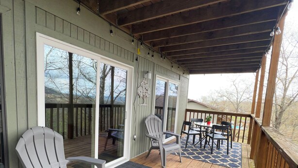 A wooden deck with two Adirondack chairs and a table, a covered area with a wooden ceiling, and a view of trees and a house in the background. - Modern treehouse in the woods near SDC! (Reeds Spring)