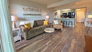 A living room with a sofa, coffee table, and a chair with a pillow that says "ENJOY TODAY". There is a painting on the wall and a kitchen area in the background. - Modern treehouse in the woods near SDC! (Reeds Spring)