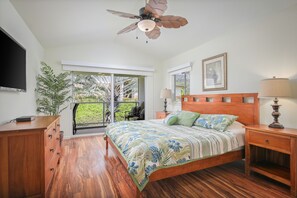 Un dormitorio con cama, mesitas de noche de madera, ventilador de techo, televisor y vistas al exterior.