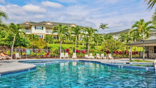 Une piscine entourée de chaises longues et de palmiers, elle-même bordée d’un jardin aménagé et d’un bâtiment doté de balcons.