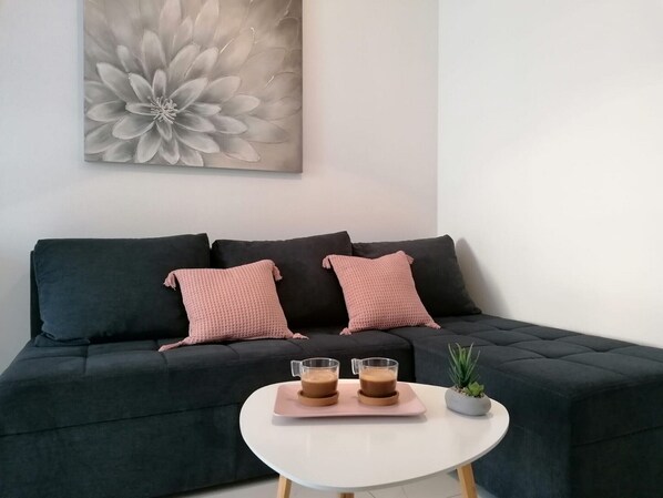 A modern living room with a dark sofa, two pink cushions, a white coffee table, and a large floral painting on the wall.