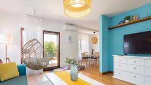 Ein modernes Wohnzimmer mit einer Couch, einem Hängekorbstuhl, einem Couchtisch mit einer Vase, einem Fernseher auf einem weißen Schrank und einem Essbereich im Hintergrund.