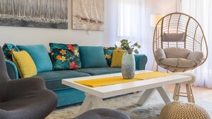 Ein modernes Wohnzimmer mit einem türkisfarbenen Sofa, bunten Kissen, einem weißen Couchtisch, einem Hängesessel und abstrakter Wandkunst.