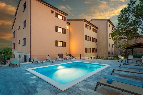 Una piscina rectangular con sillones de descanso y un edificio con múltiples ventanas y balcones.