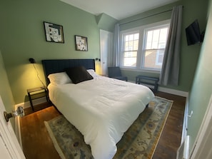 A bedroom with a large bed, a nightstand, a chair, a small table, and a window with curtains. - 5 minutes from major highways, close to EVERYTHNIG! (Parma)