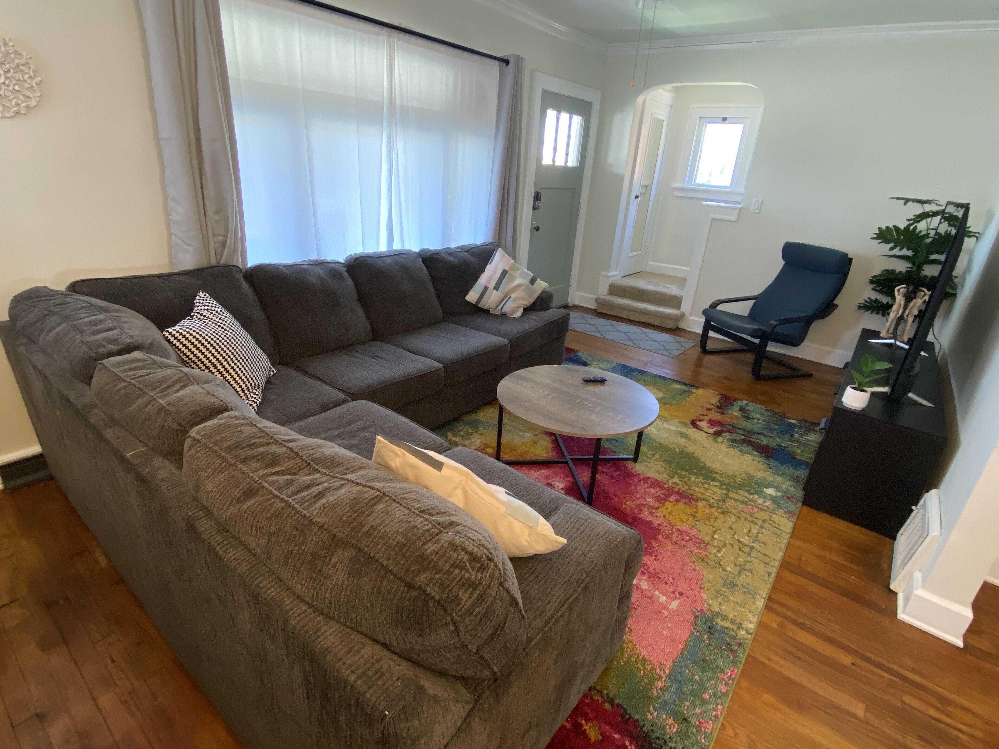 Ein Wohnzimmer mit einem grauen Ecksofa, einem kleinen runden Couchtisch, einem blauen Sessel und einem farbenfrohen Teppich.