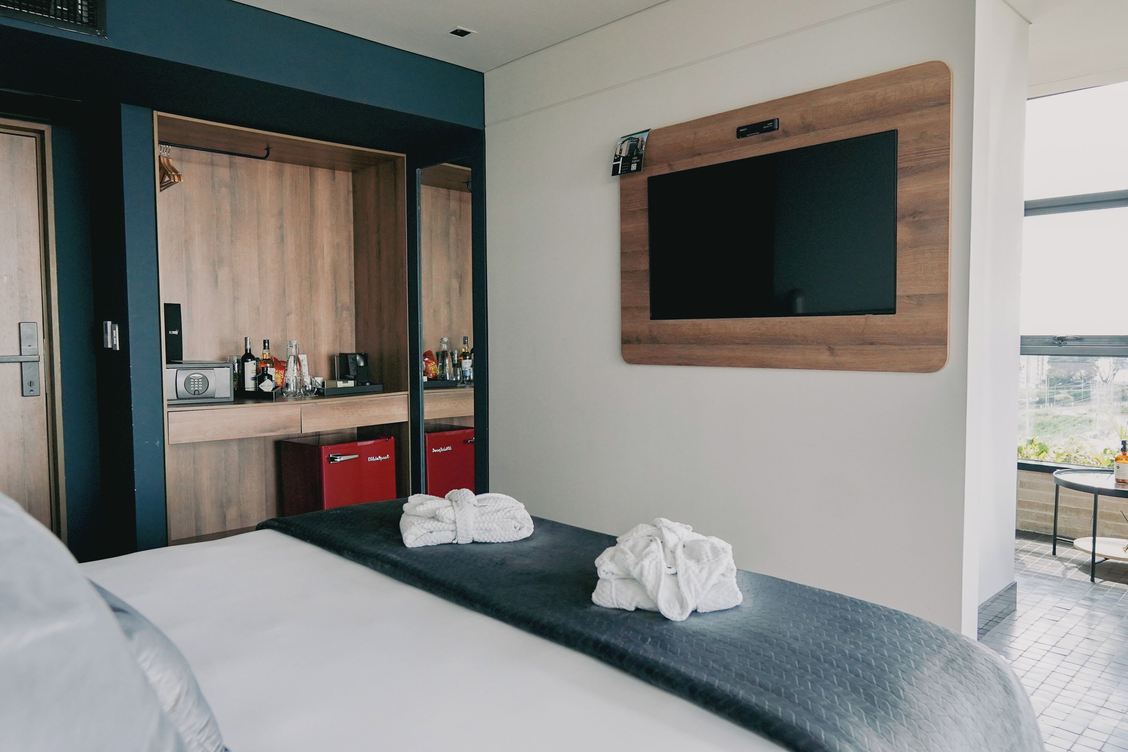 Habitación de hotel con cama, espejo, televisor montado en la pared y una pequeña mesa con una silla.