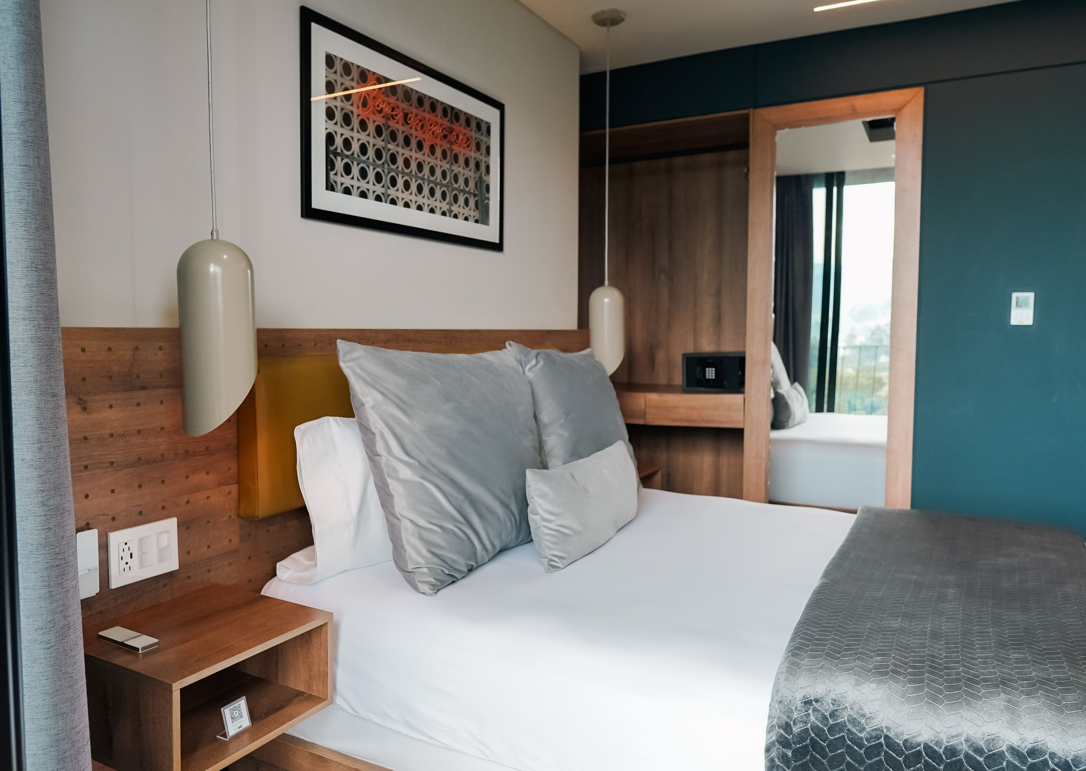 Ein Hotelzimmer mit Bett, Holz-Kopfstütze, Nachttisch, Lampe, einem gerahmten Bild an der Wand und einem Fenster mit Aussicht.